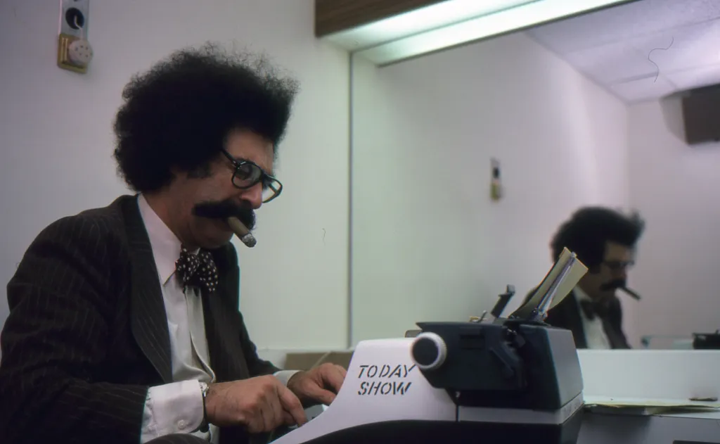 Journalist Gene Shalit smoking a cigar while typing on a typewriter labeled "Today Show" at NBC Studios.