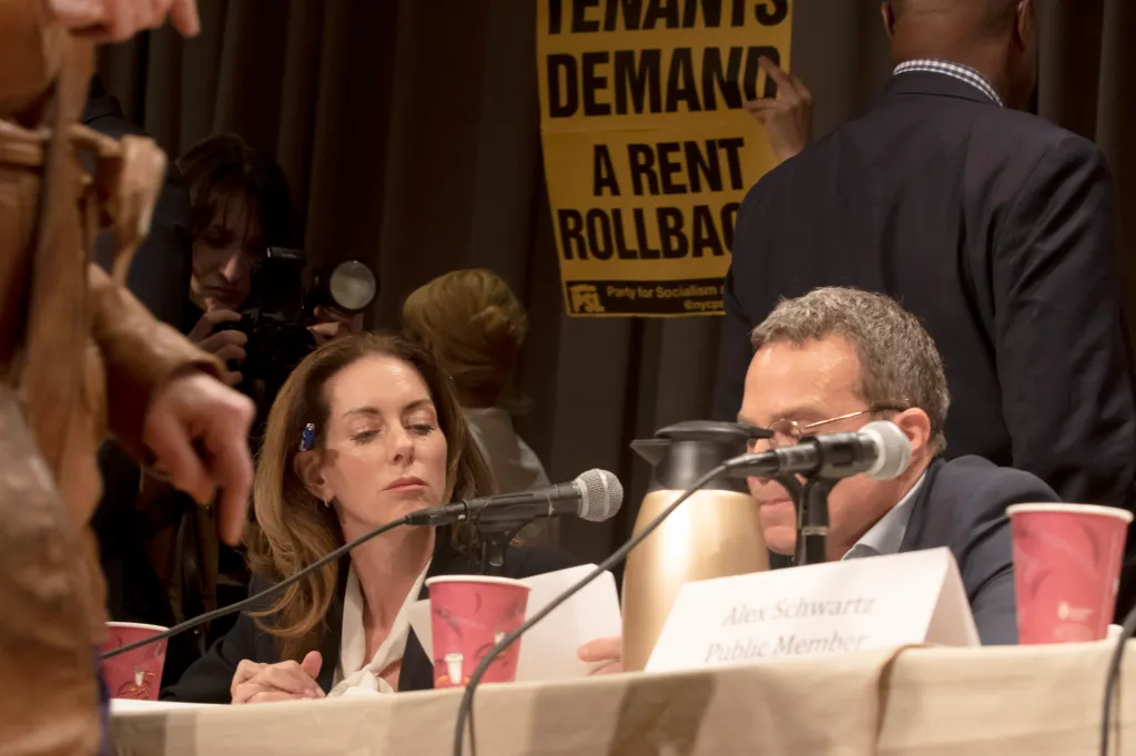 Landlord representatives Christina Smyth and Robert Erlich at a Rent Guidelines Board meeting, with a protest sign "Tenants Demand A Rent Rollback" in the background.
