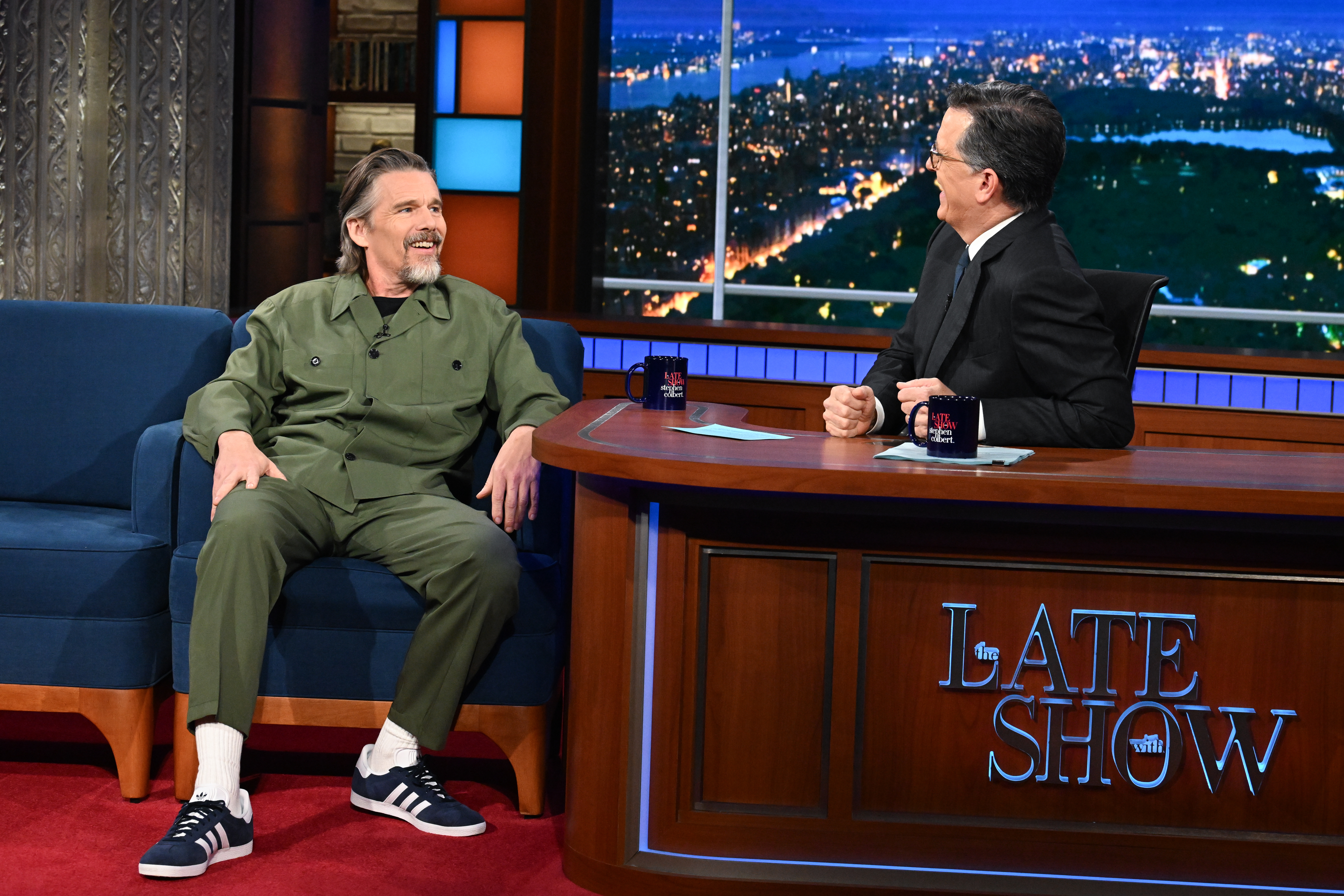 Ethan Hawke sitting on a blue couch across from Stephen Colbert at his desk during "The Late Show."