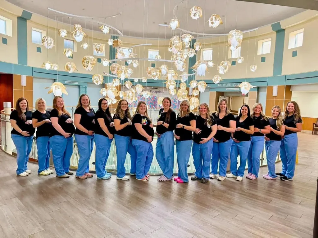 A group of 17 pregnant nurses standing in blue scrubs and black shirts, cradling their baby bumps.