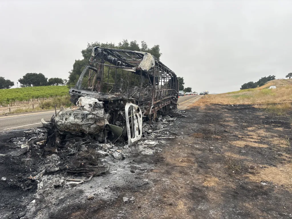 A burned-out bus wreck on the shoulder of a highway after a collision.