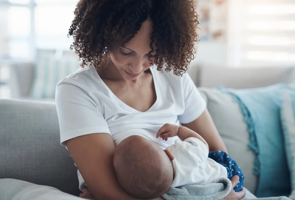 A mother with curly hair breastfeeds her baby on a couch.