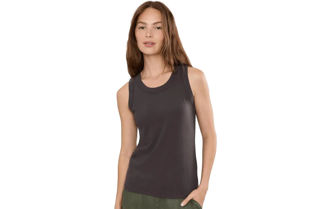 A young woman with brown hair and light skin, wearing a dark gray tank top and green pants, stands facing forward with her right hand in her pocket.