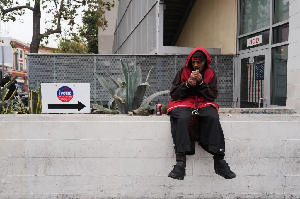 A man in a red hoodie lighting a cigarette outside a voting center with an "I Voted" sign nearby.