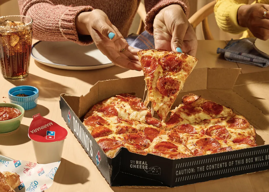 A person taking a slice of pepperoni pizza from a Domino's box.