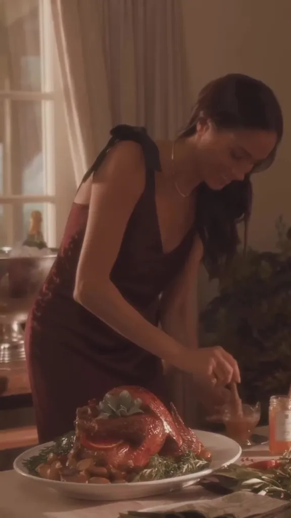 Meghan Markle in a maroon slip dress preparing a roasted turkey.