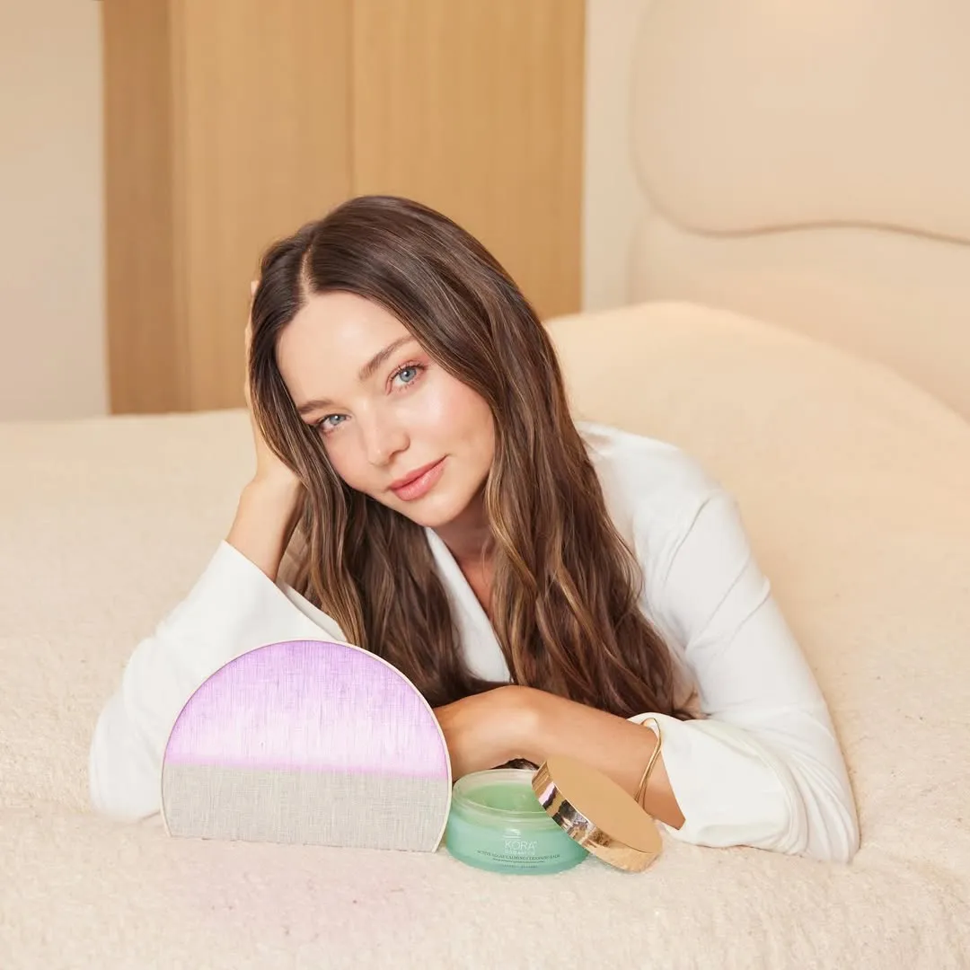 Miranda Kerr reclines on a bed, next to a pink-and-white vanity mirror and an open jar of KORA Organics face cream.