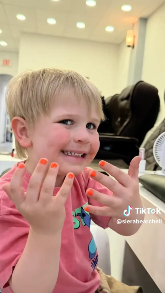 A smiling child with blonde hair showing off bright orange painted fingernails.