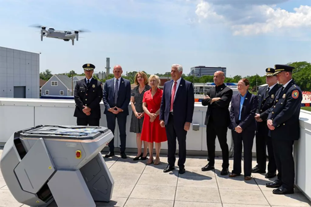 Nassau County Executive Bruce Blakeman and other officials stand on a rooftop next to a drone first responder program base station, as a drone flies overhead.