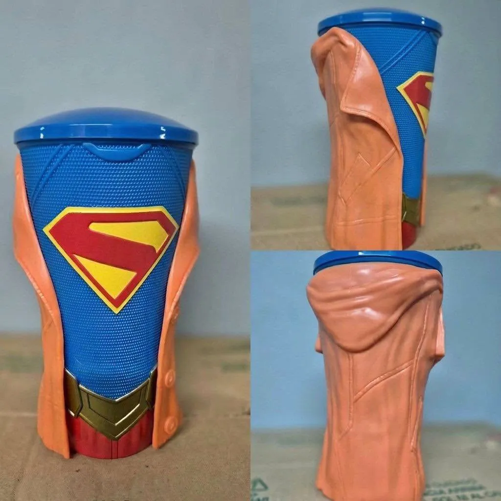 A Supergirl movie drink cup with a blue cup, Superman logo, gold belt buckle, and orange jacket.