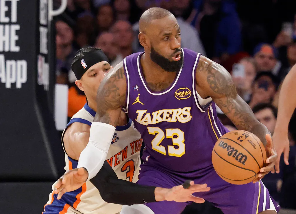 LeBron James, in a purple Lakers jersey, dribbles a basketball while being defended by Josh Hart of the Knicks.