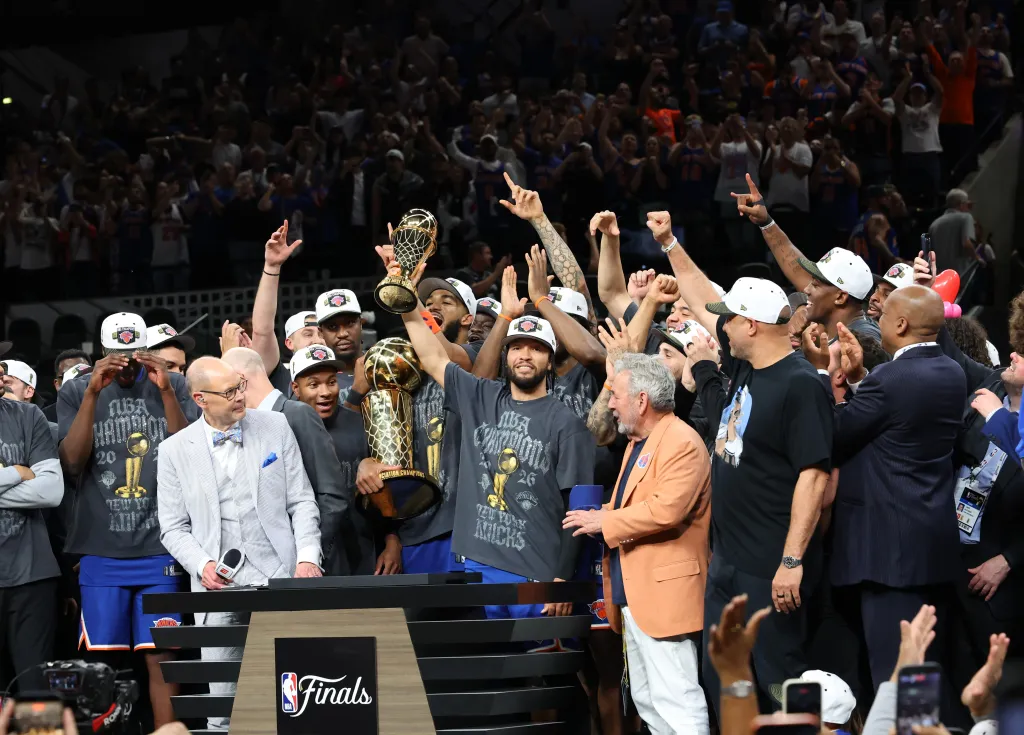 The New York Knicks celebrate after winning the 2026 NBA Finals against the San Antonio Spurs, holding up championship trophies.