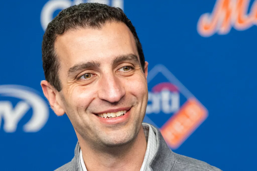 David Stearns, President of Baseball Operations for the New York Mets, smiling against a blue background with blurred logos.