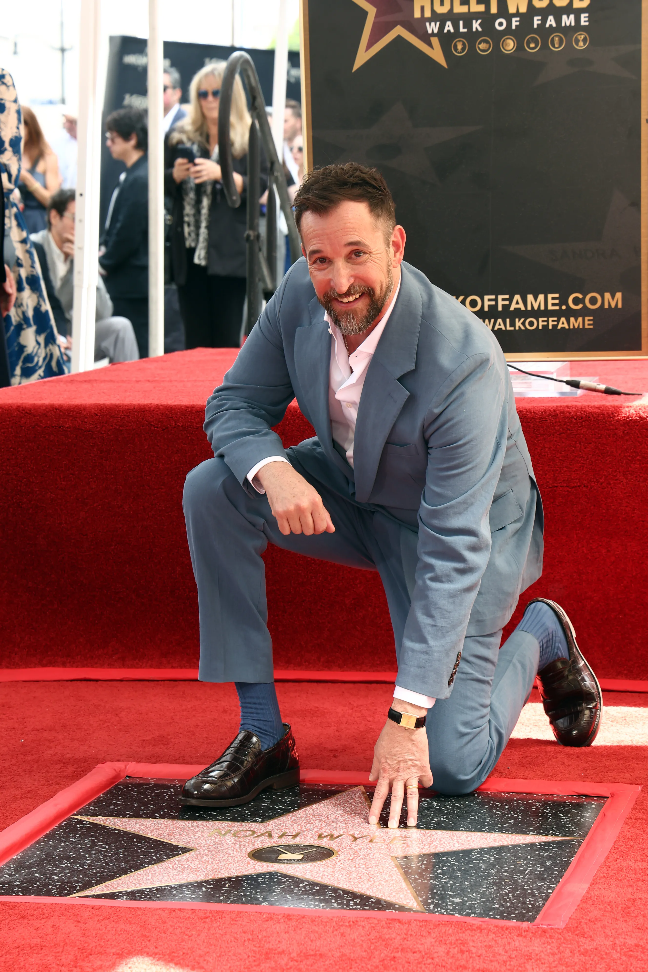 Noah Wyle kneeling on a red carpet next to his newly unveiled star on the Hollywood Walk of Fame.