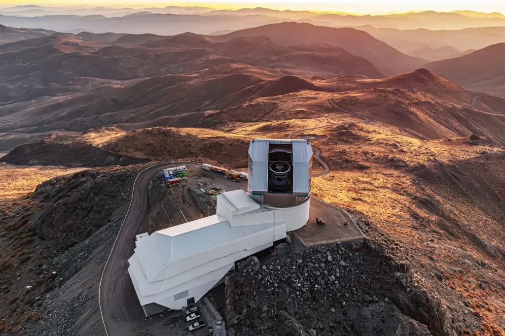 Aerial view of the Vera C. Rubin Observatory perched on a mountain, with a vast mountain range bathed in golden light in the background.