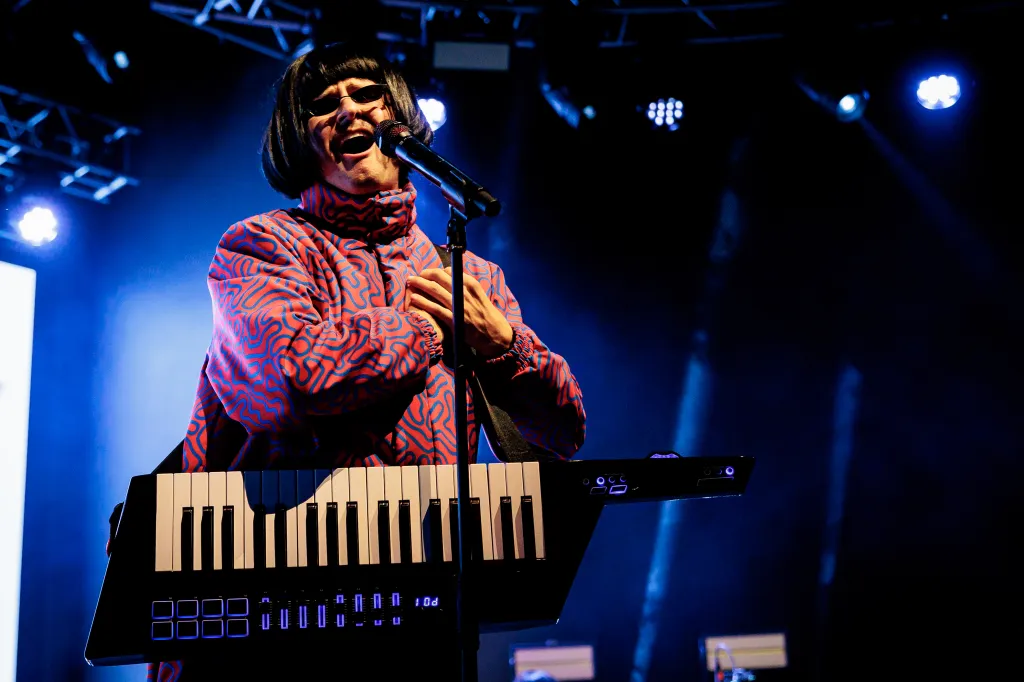 Oliver Tree performing on stage, wearing a red and blue patterned jacket and black wig, singing into a microphone, with a keyboard in front of him.