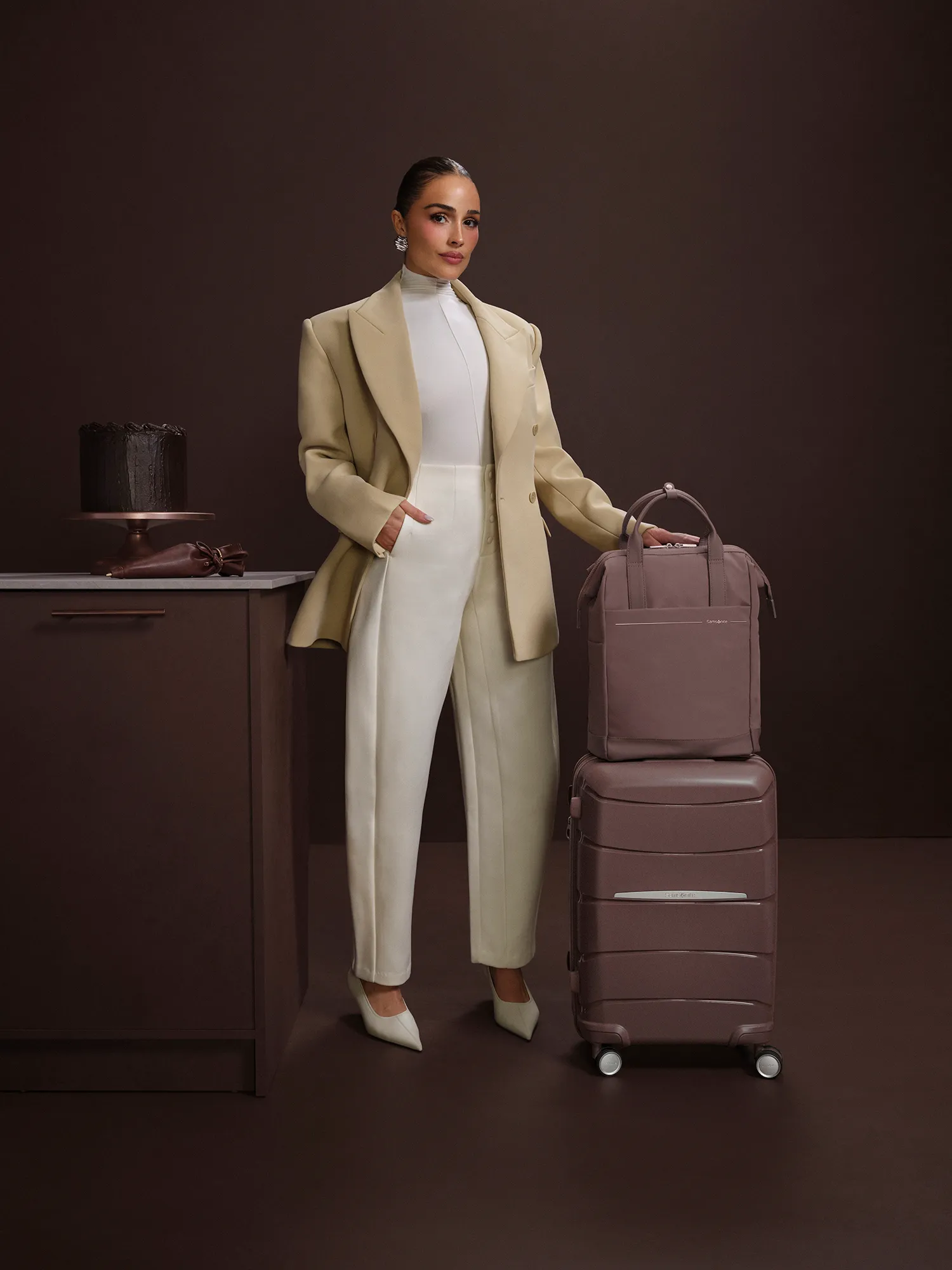 Olivia Culpo stands next to Samsonite luggage and a chocolate cake.