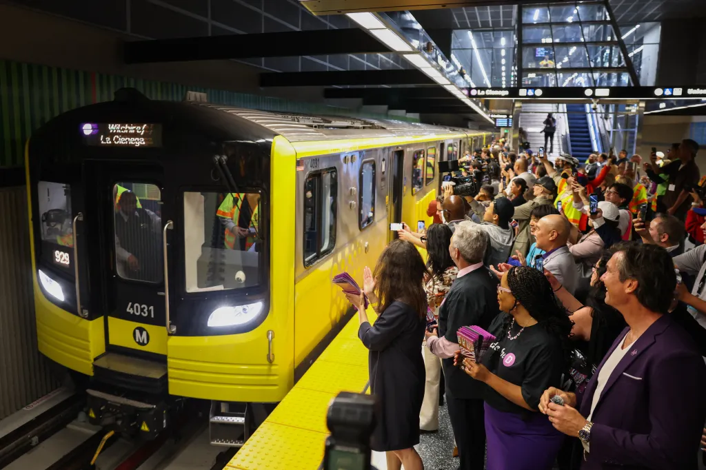 A new Metro D Line subway train arrives at the Wilshire/Fairfax station as people on the platform cheer and take photos.
