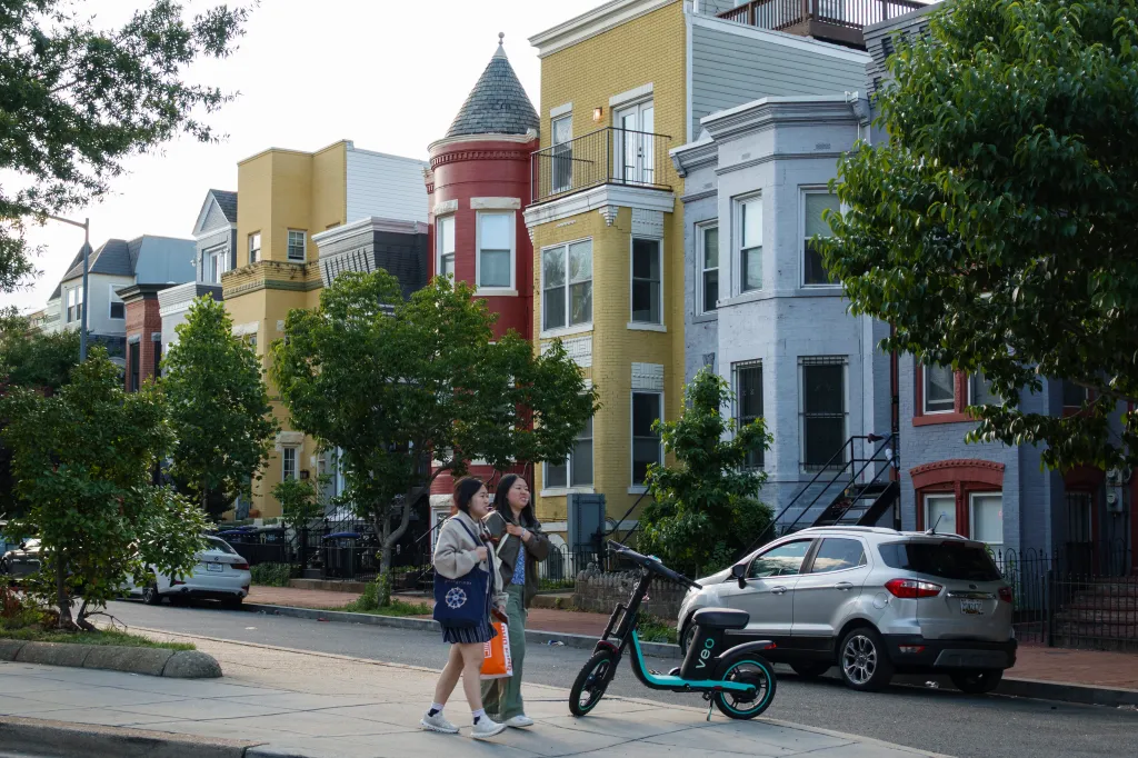 Two women walking past colorful row houses and a scooter parked on the sidewalk.