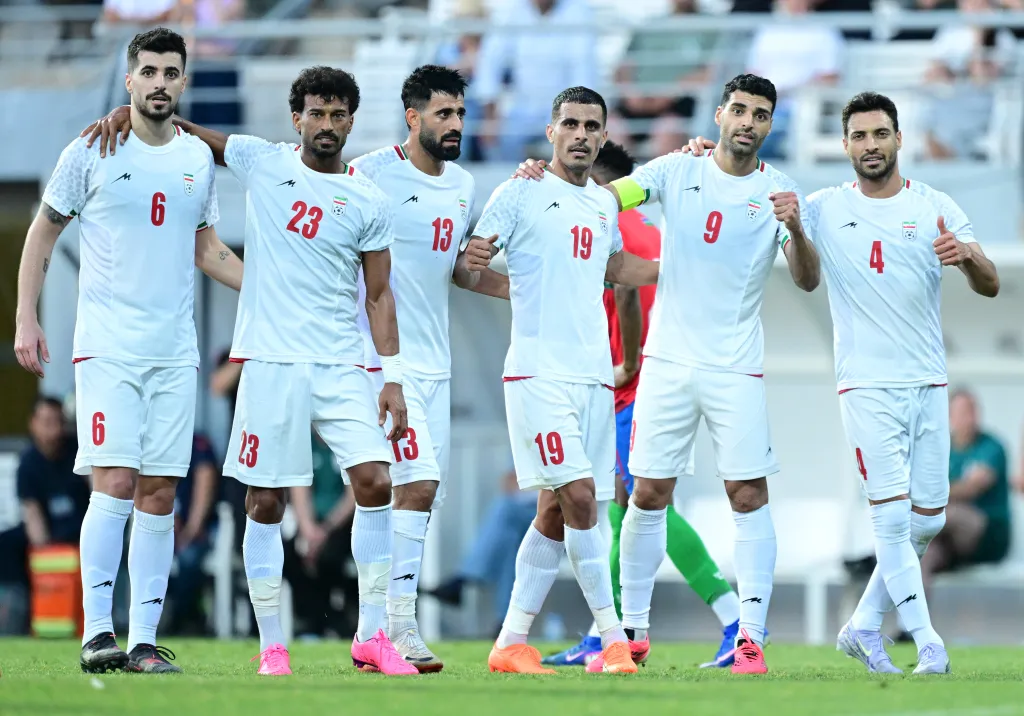 Six Iranian national football players in white jerseys celebrating on the field.