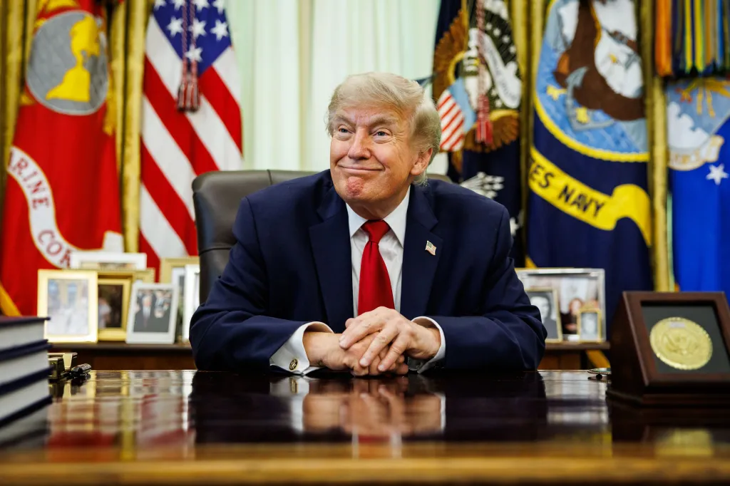 President Donald Trump smiles while seated at a desk in front of several flags in the Oval Office.