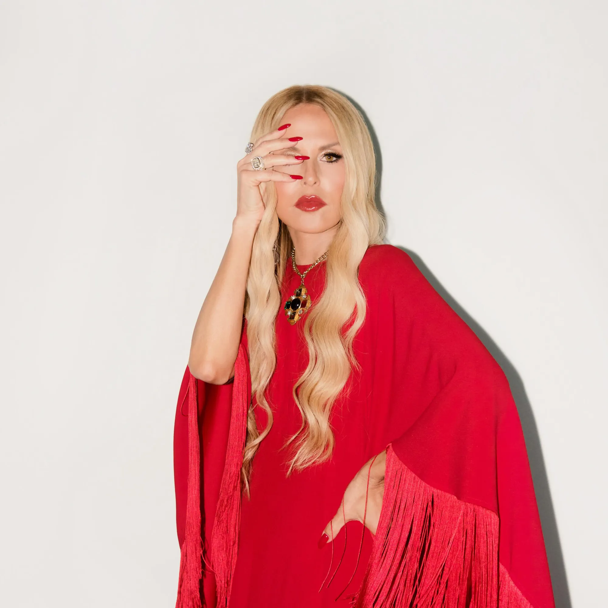 Rachel Zoe in a red dress with a fringed sleeve, covering part of her face with one hand.