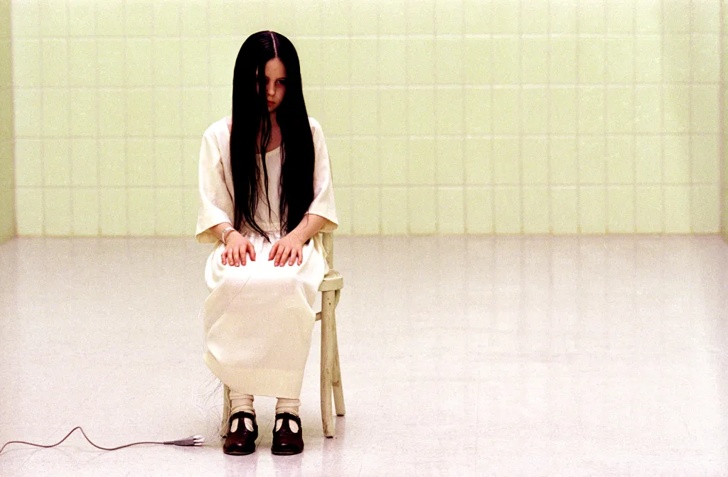 Daveigh Chase as Samara in The Ring (2002), sitting on a chair with a cable attached to her.
