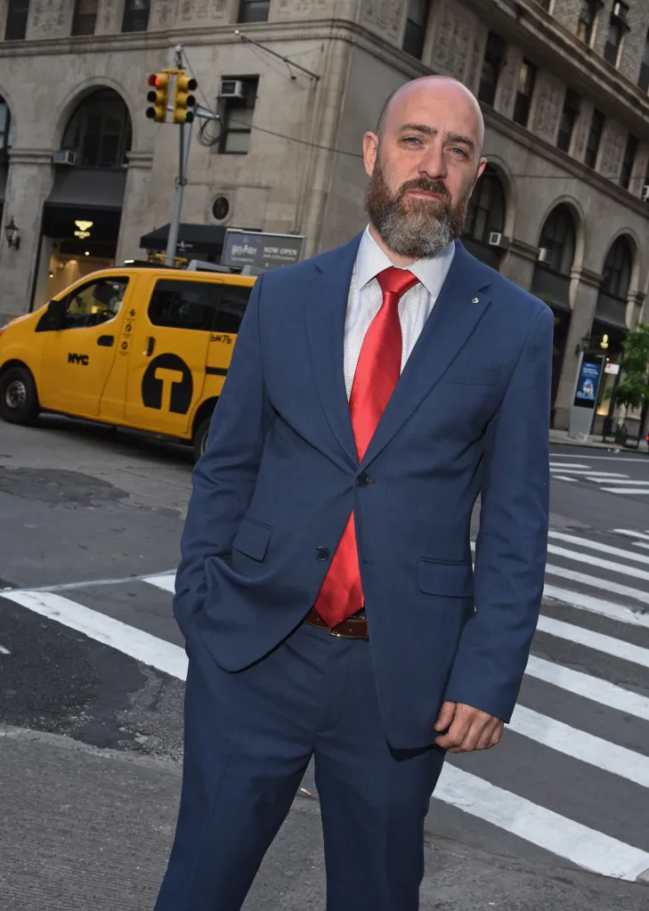 Jonathan David Rinaldi, a bald man with a beard, in a blue suit and red tie, stands in a crosswalk in New York City with a yellow taxi behind him.
