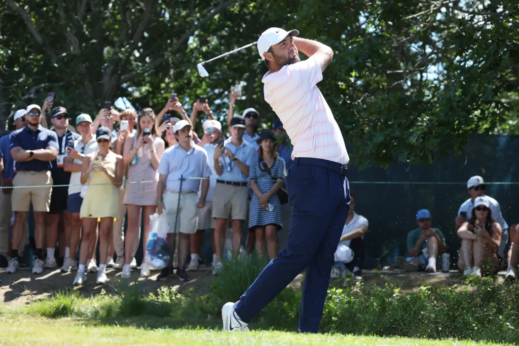 Scottie Scheffler tees off, facing left with a golf club swung overhead, as spectators watch behind him.
