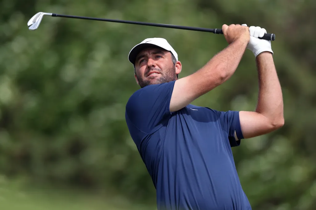 Scottie Scheffler in a blue shirt and white hat, mid-swing with a golf club.