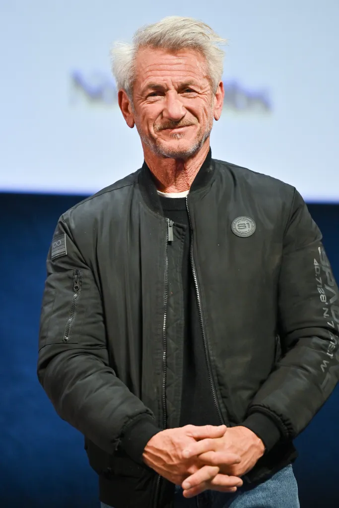 Sean Penn smiling while attending an event in a black jacket and blue jeans.
