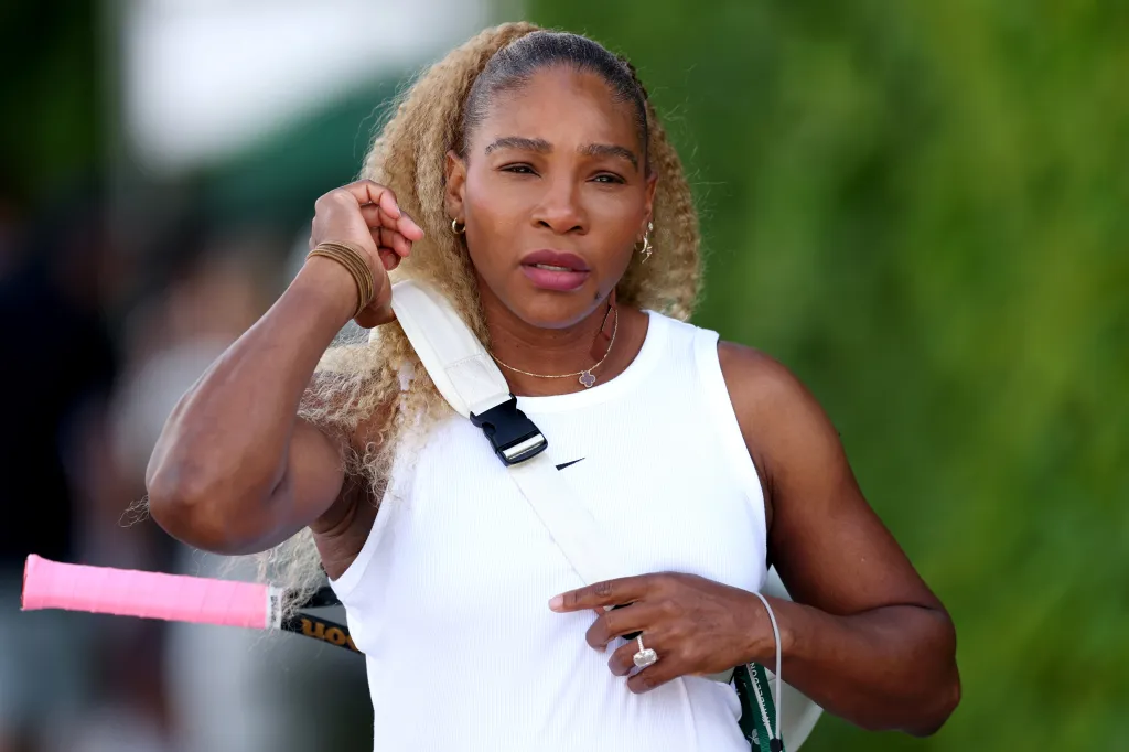 Serena Williams walks onto the training grounds at Wimbledon, carrying a tennis racket and wearing a white ribbed tank top.