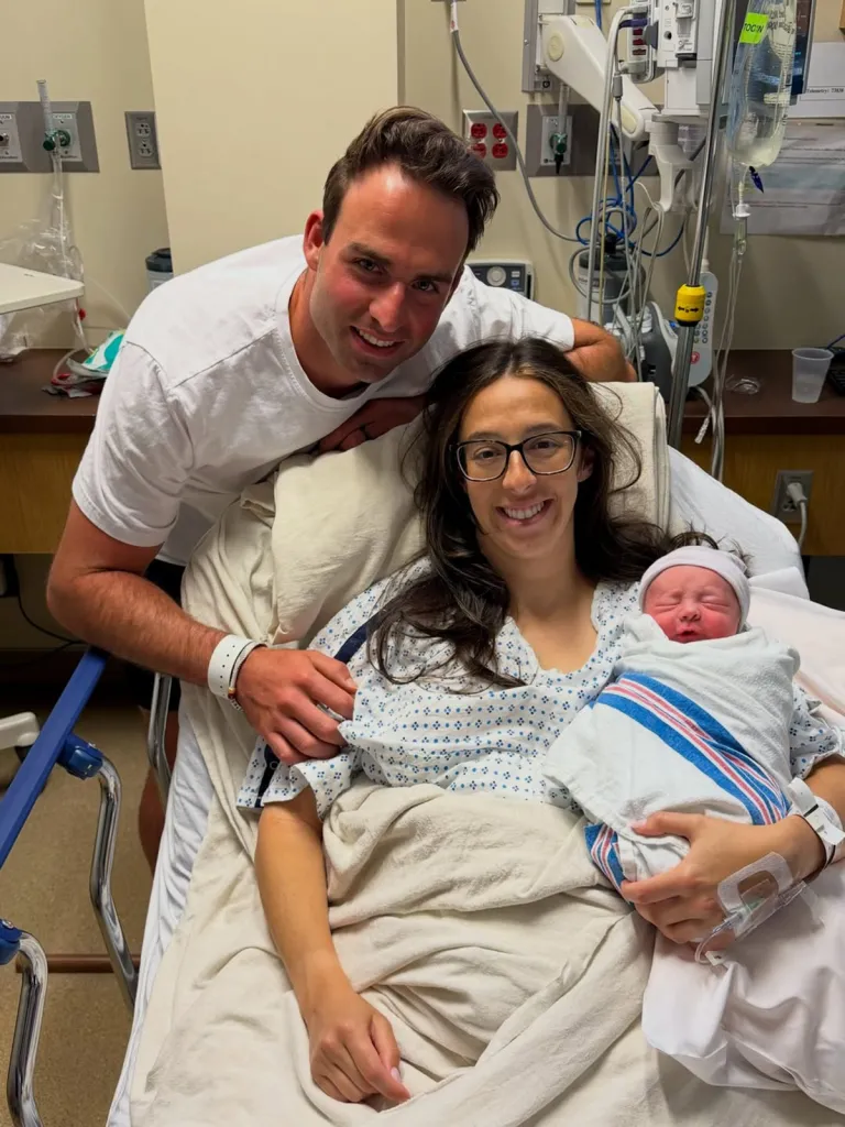 A man stands behind a woman in a hospital bed holding a swaddled newborn, all three smiling.