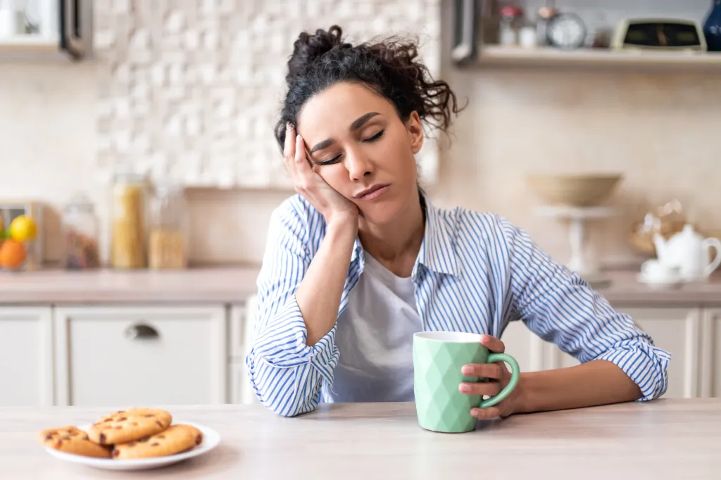 Sleepy young woman with closed eyes holding a cup at a kitchen table.