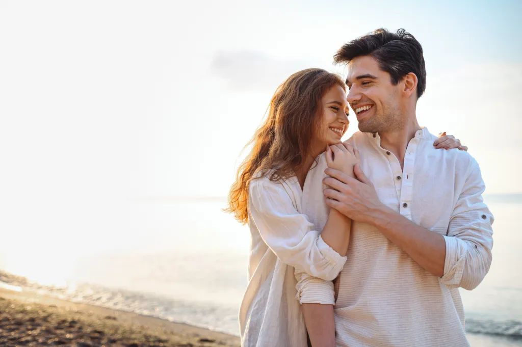 Smiling couple embracing on a beach with the ocean in the background.