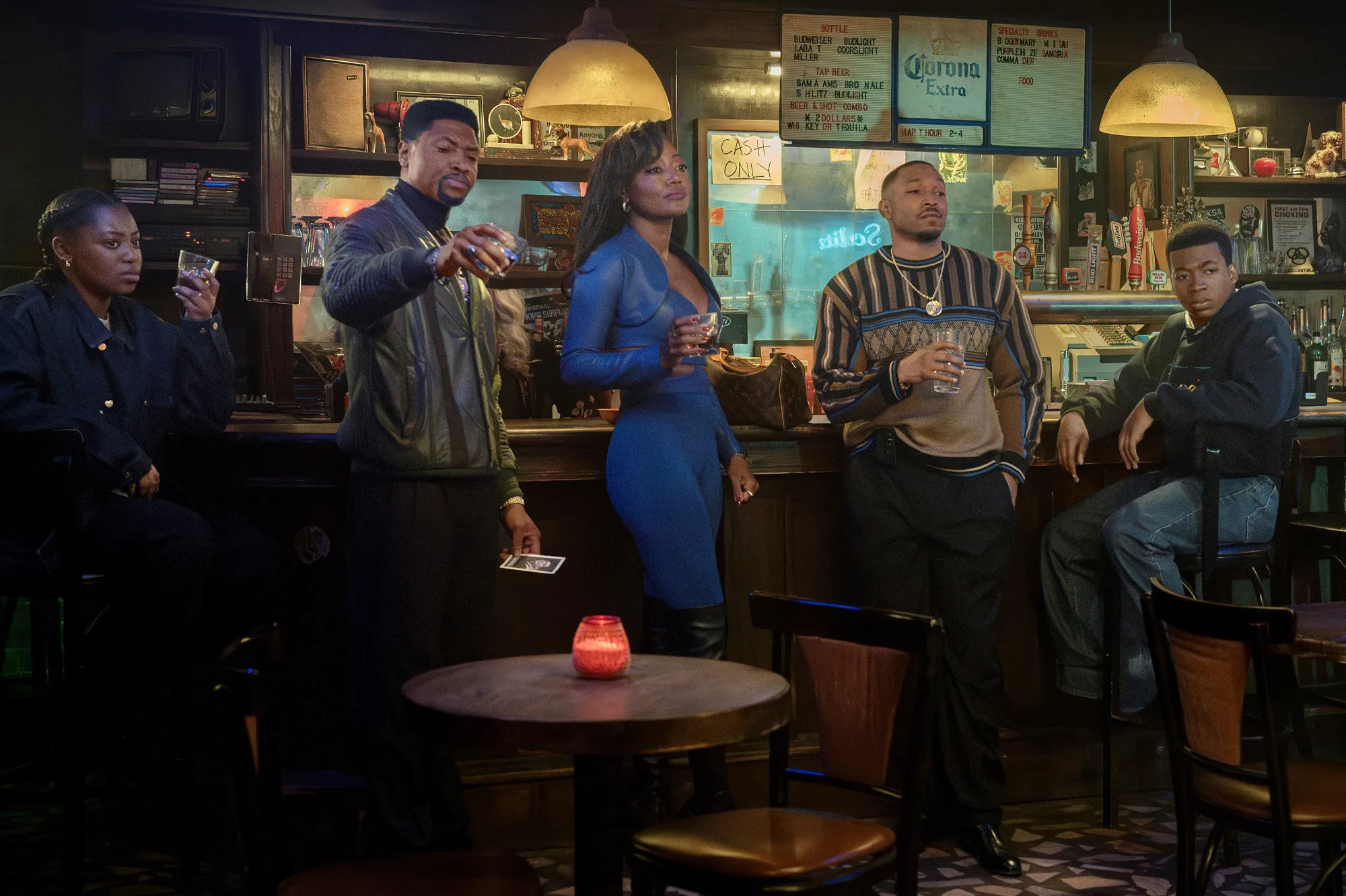 Five people standing and sitting at a bar with drinks.