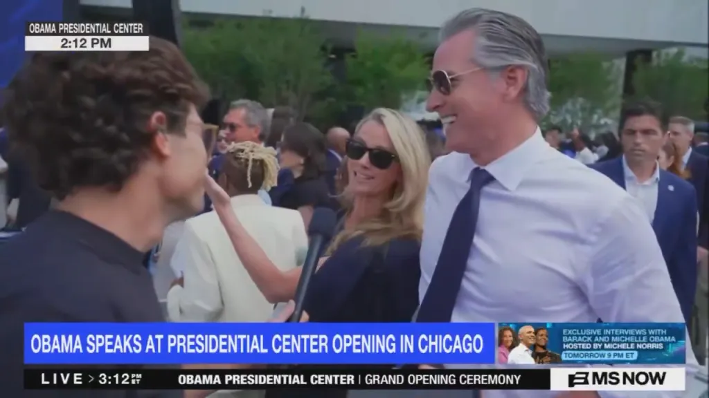 Gavin Newsom smiling at a reporter holding a microphone, with a crowd of people in the background.