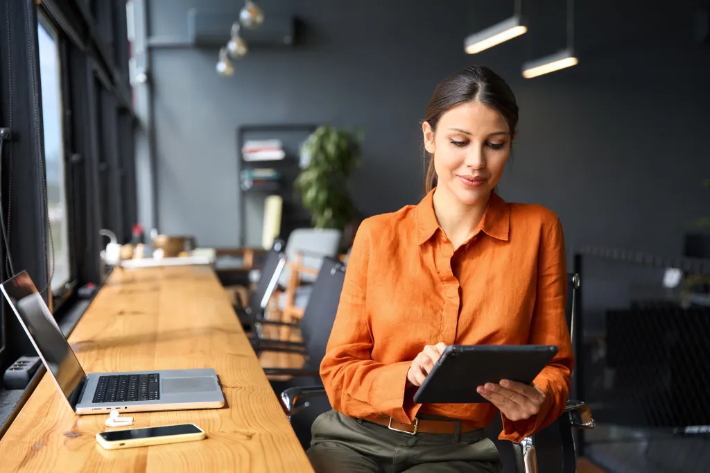 A young Hispanic woman sitting at a desk and using a tablet in a modern office.
