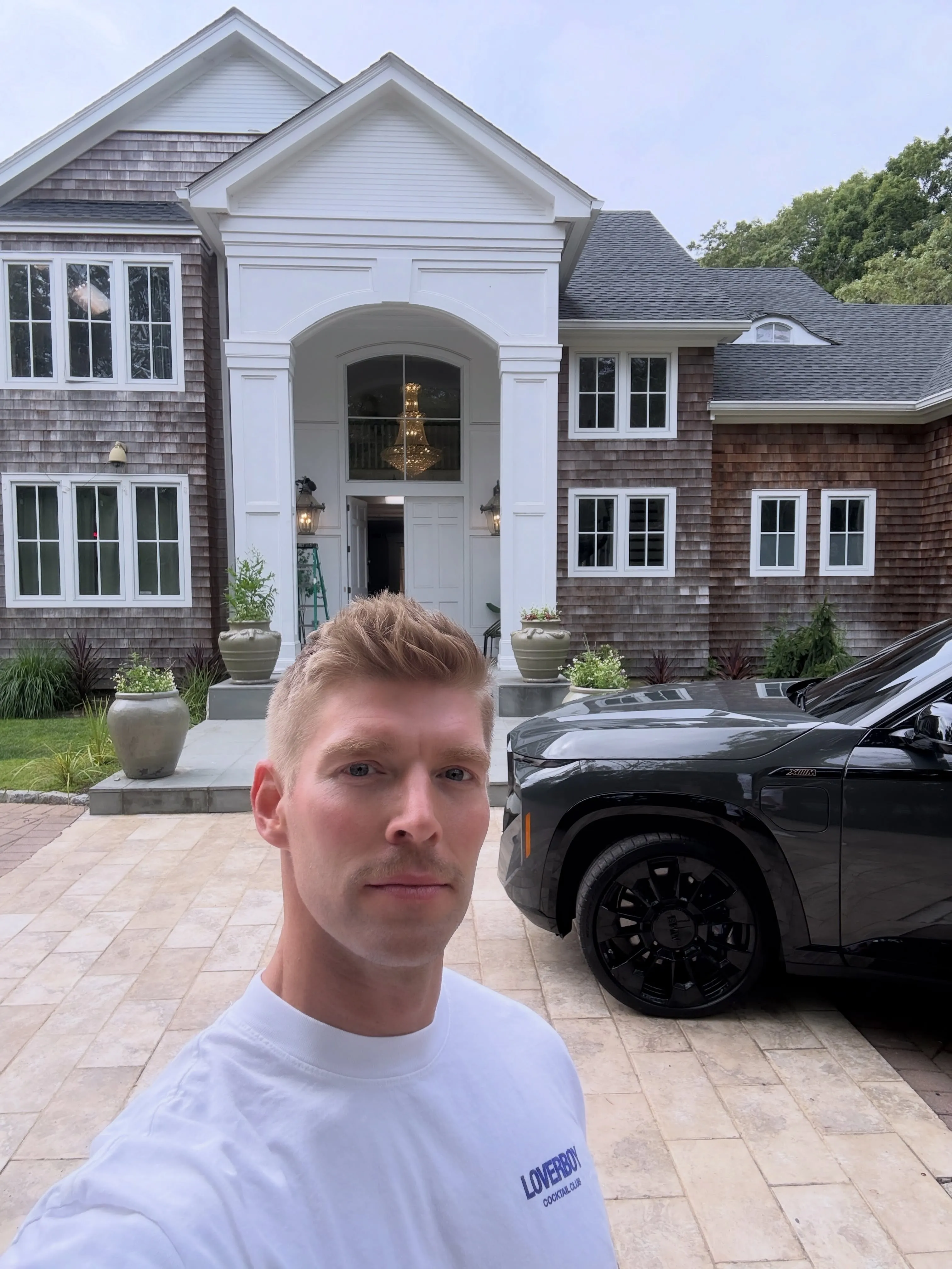 Kyle Cooke poses in front of a large house and a black car.