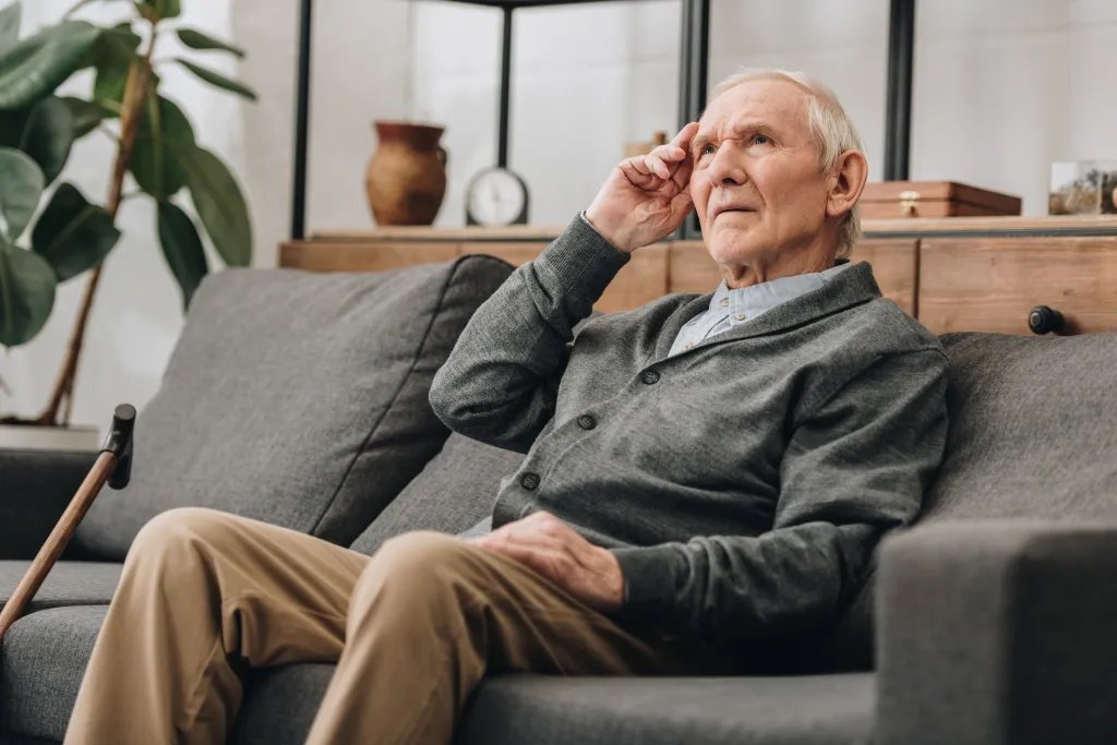 A thoughtful senior man with grey hair sits on a sofa in a living room, looking upwards with his hand on his forehead.