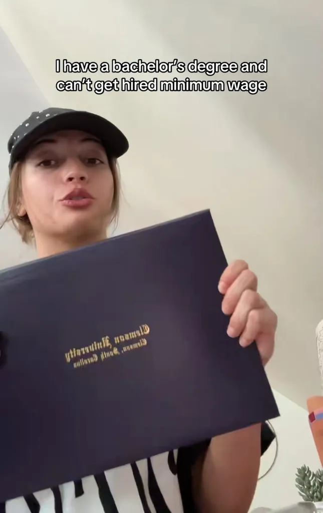 A woman wearing a baseball cap holds up her Clemson University diploma, with text overlay: "I have a bachelor's degree and can't get hired minimum wage."