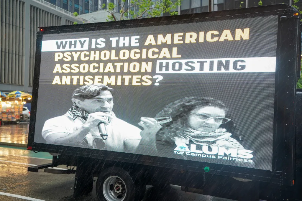 A digital billboard on the side of a truck asks, "Why is the American Psychological Association hosting antisemites?" under a black and white image of two individuals.