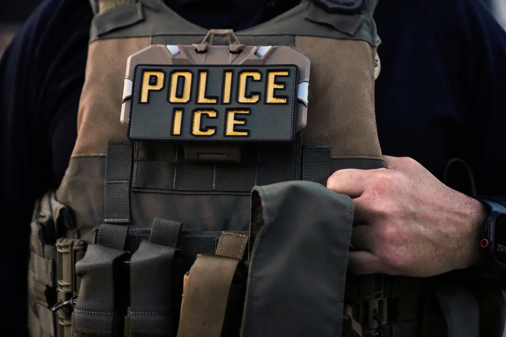 Close-up of an Immigration and Customs Enforcement (ICE) agent wearing a tactical vest with a "POLICE ICE" patch on the chest.