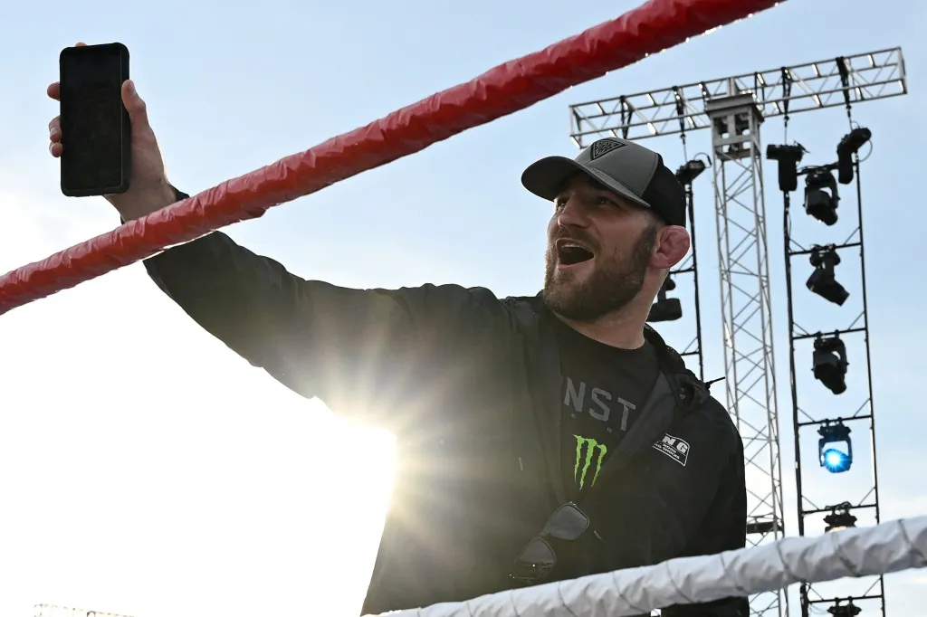 UFC Middleweight Champion Sean Strickland reacts with an open mouth while holding up a phone, with a red wrestling ring rope and bright sky behind him.
