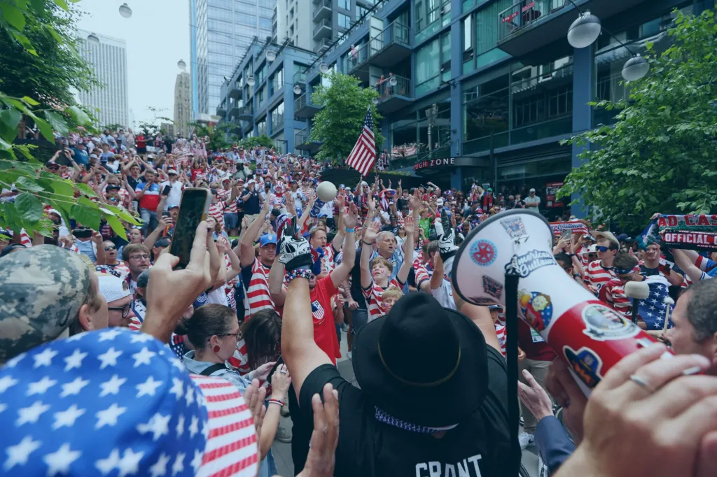 An image collage containing 1 images, Image 1 shows Crowds of fans dressed in red, white, and blue, wave their hands in the air and cheer for the US soccer team in Seattle