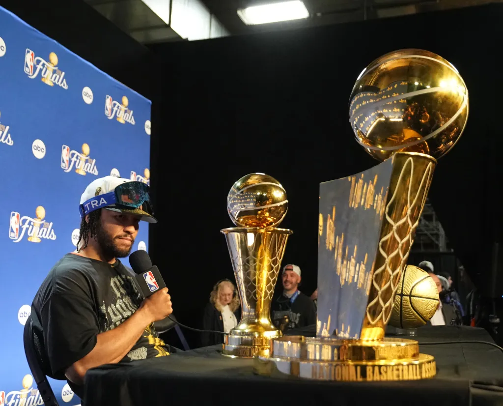 Jalen Brunson at a press conference with the Larry O'Brien Championship Trophy and a smaller trophy to his right.