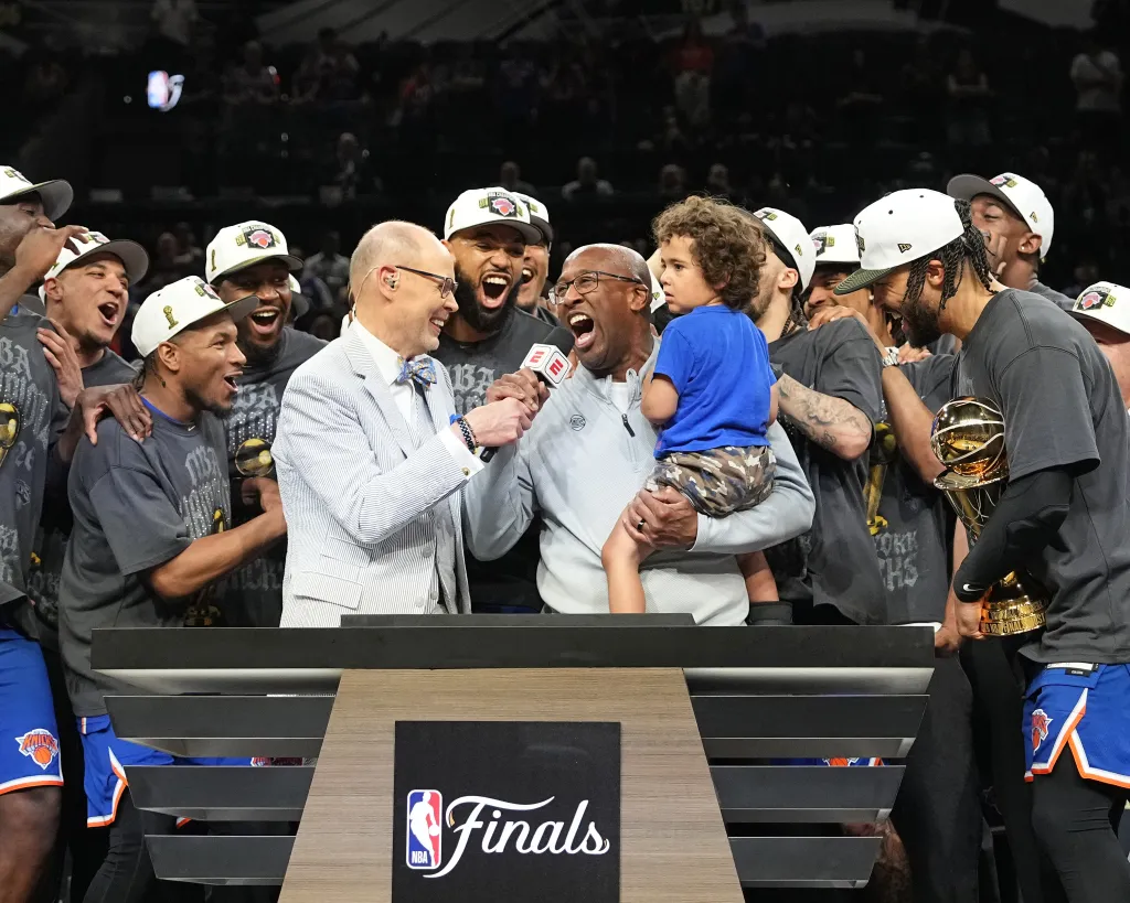 New York Knicks Head Coach Mike Brown smiles, holding a child and an NBA Finals trophy, while being interviewed amidst celebrating players wearing championship hats.