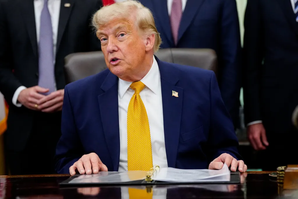 Donald Trump seated at a desk, speaking, with an open binder in front of him.
