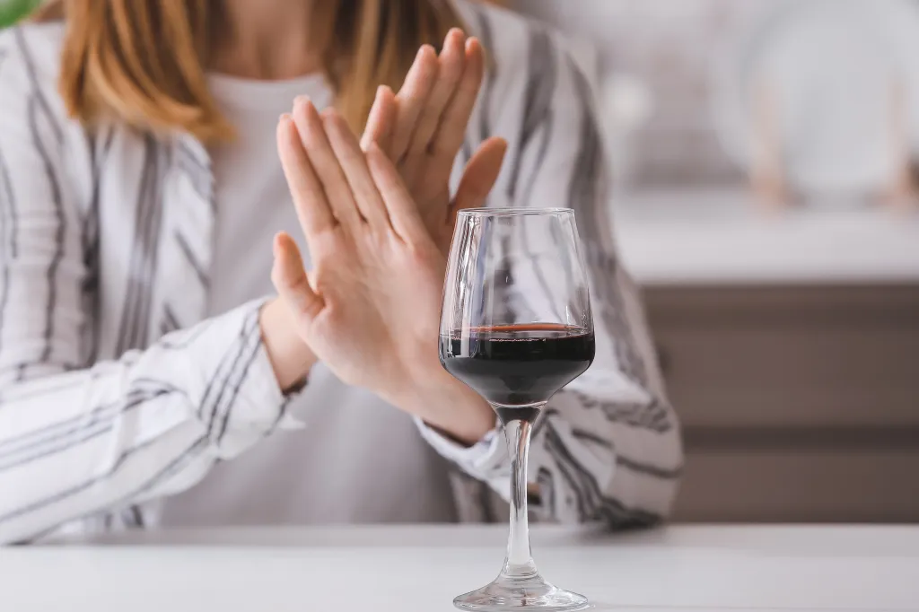 A woman in a white and black striped shirt holds her hands up in a "no" gesture in front of a glass of red wine.