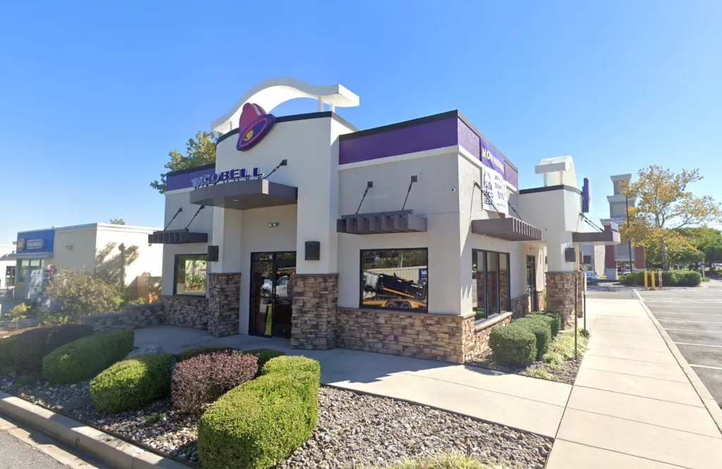 A Taco Bell restaurant building with a small parking lot, bushes, and a paved walkway.